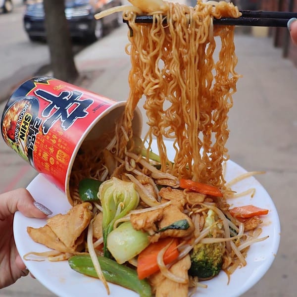 Chopsticks lift noodles from a Shin Ramen cup spilling onto a plate of stir-fried vegetables and tofu.